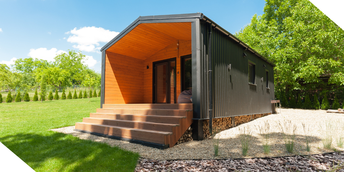 A sleek modular black and wood cabin with steps, set amidst green grass and trees, perfect as a new construction real estate investment property.