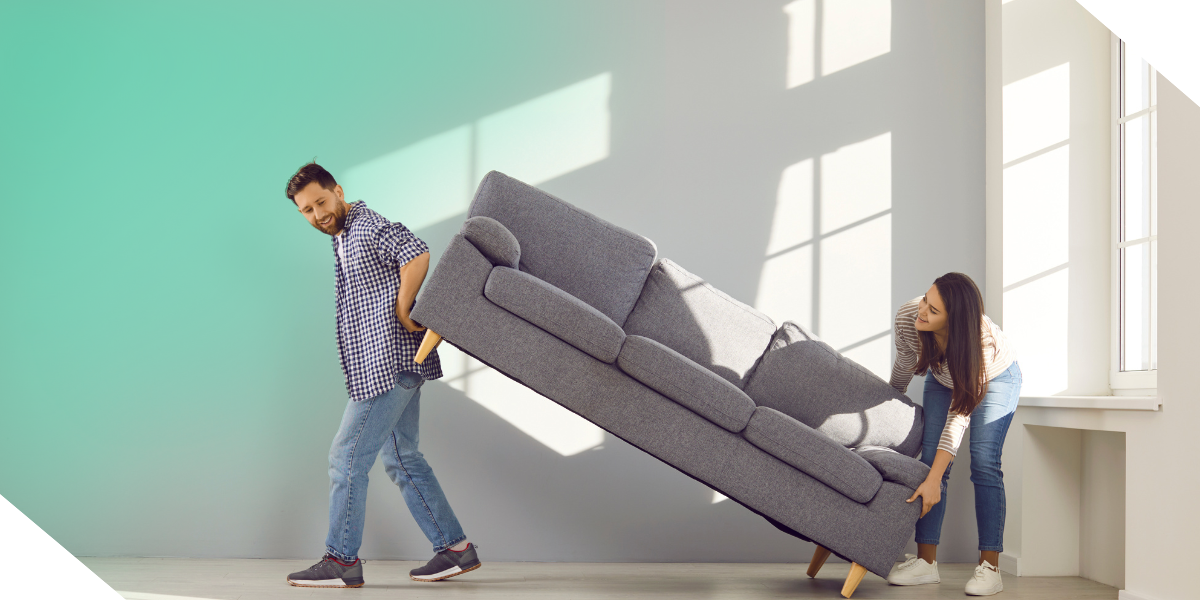 Two real estate investors move a gray couch together in a sunny room, staging the fix and flip property to be listed for sale.