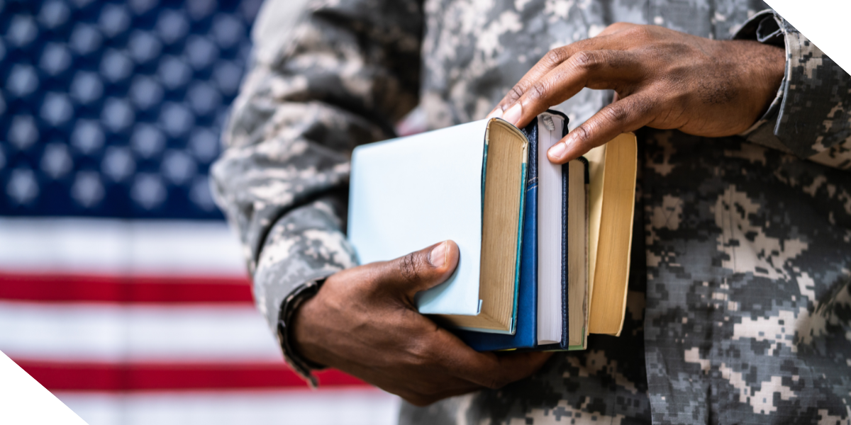 A soldier in camouflage holds a stack of books in front of an American flag. 