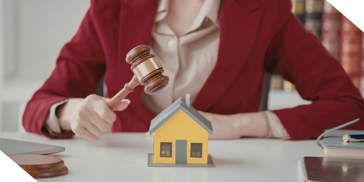 A real estate investor in a red blazer holds a gavel above a small yellow model house on a table.