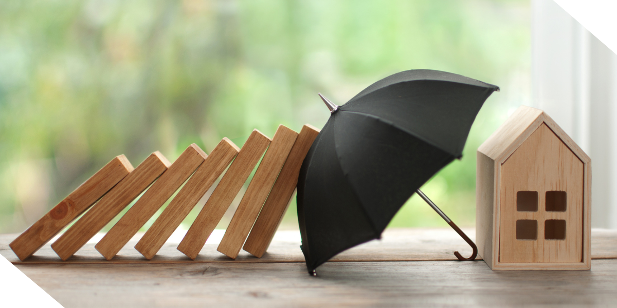 A metaphorical image of a black umbrella shielding a small wooden house and foundational wooden blocks, representing how umbrella insurance protects real estate assets.