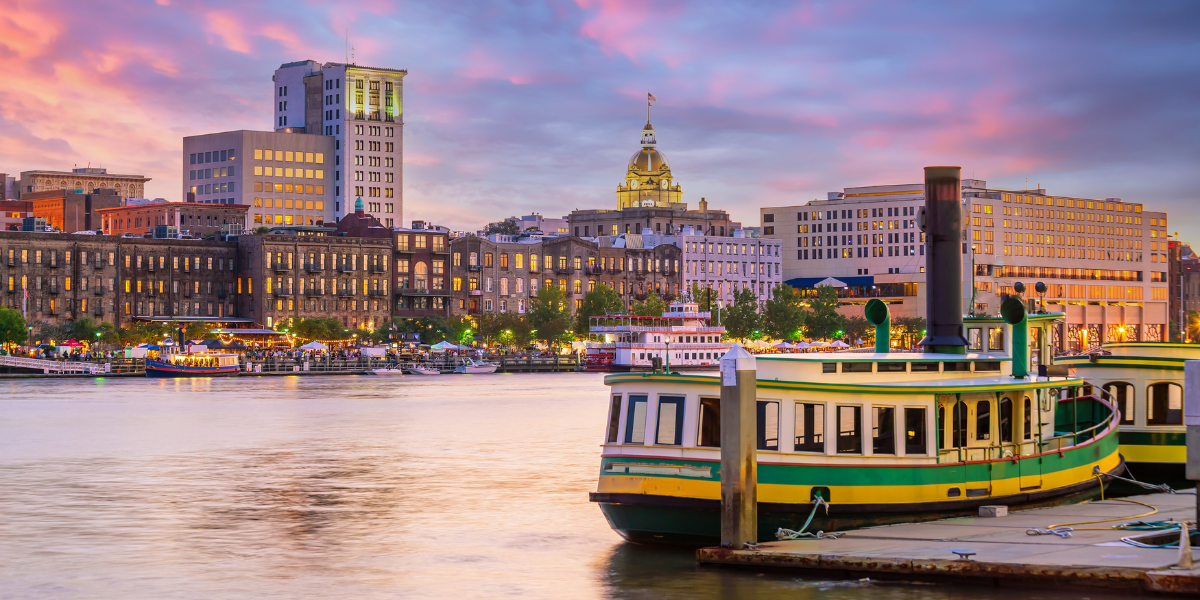 A green and yellow riverboat docked along the historic riverfront of Savannah, Georgia at dusk.