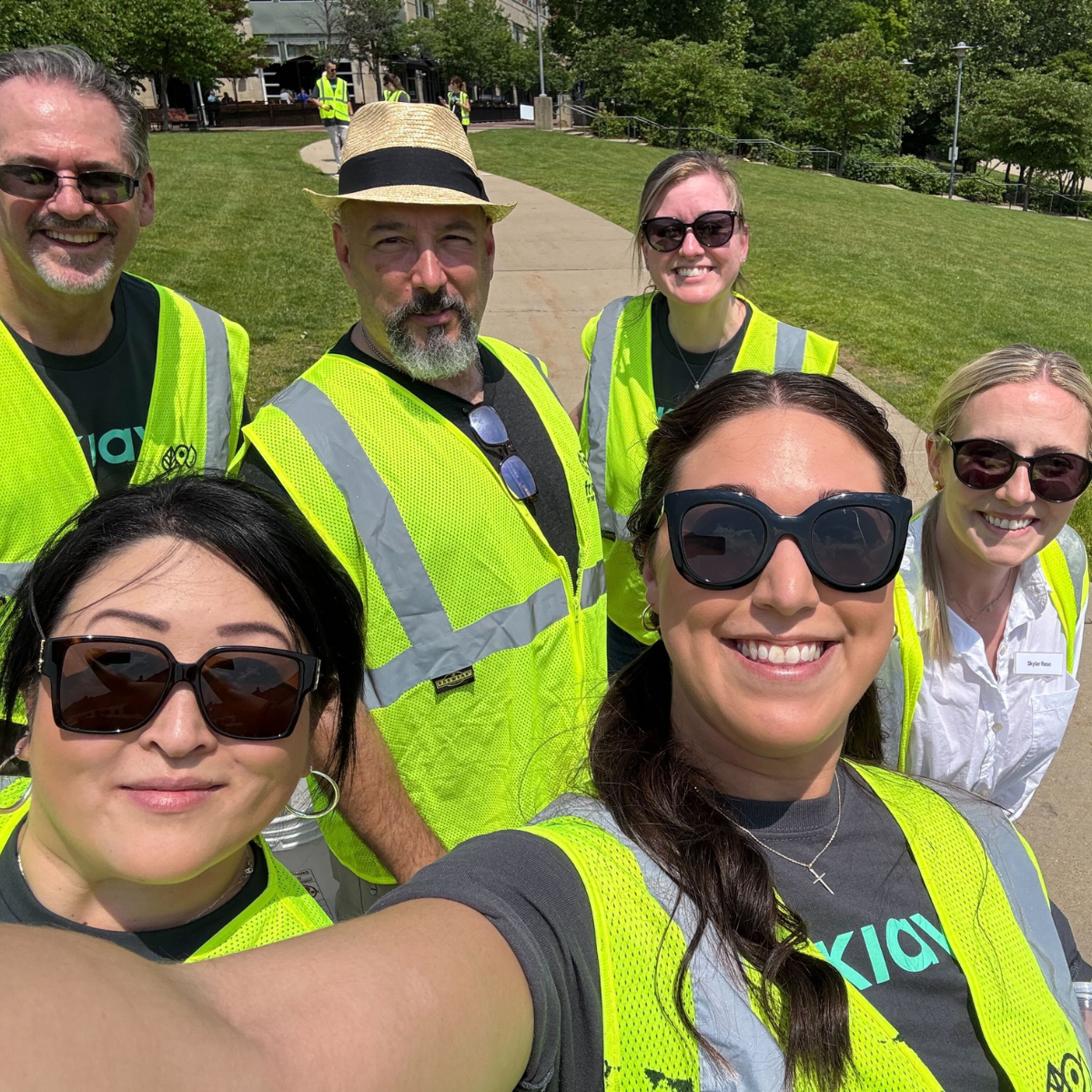 A group of six Kiavi employees wearing bright yellow safety vests posing for a selfie outdoors.
