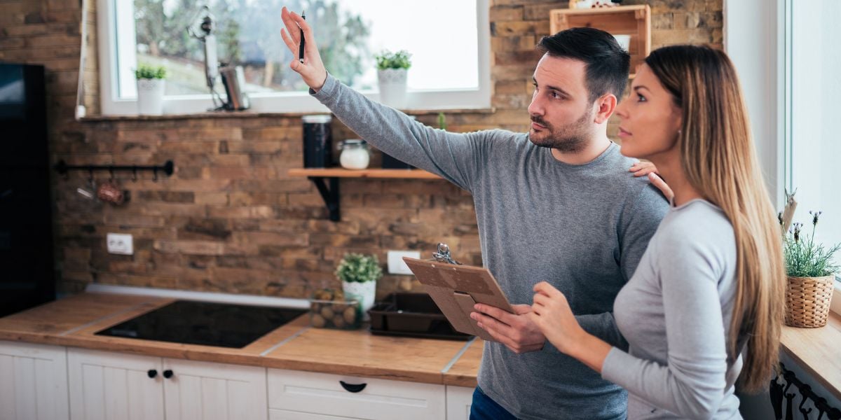 A real estate investor couple assesses a room for renovation, with the man holding a clipboard and pointing upwards towards a corner, while the woman looks on thoughtfully, suggesting a planning phase of a fix and flip project.