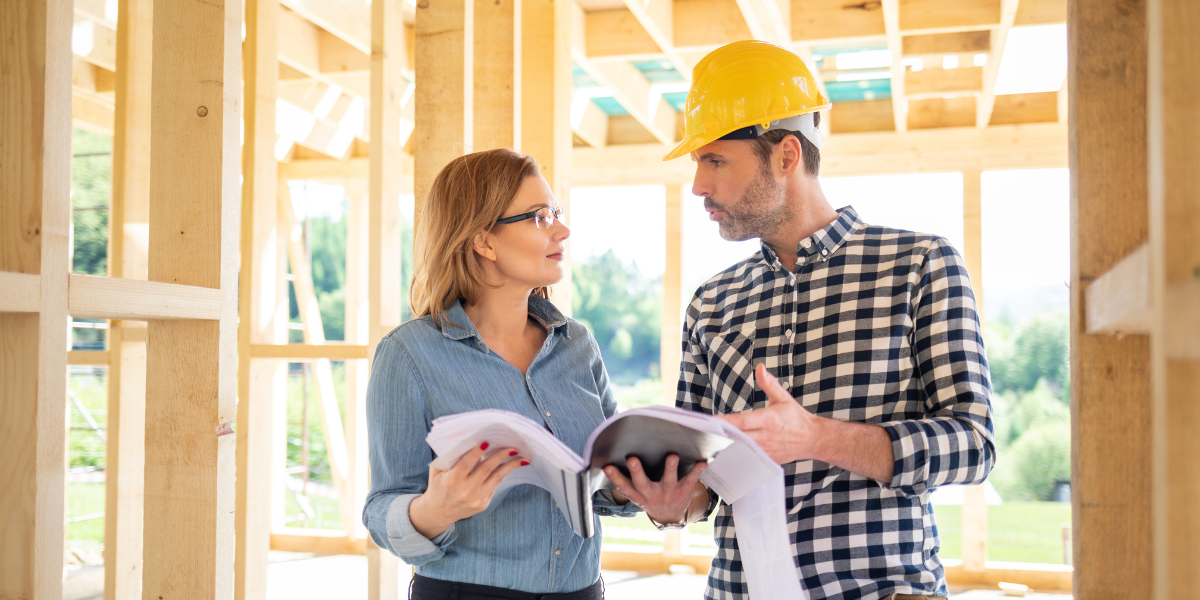 A man in a hard hat and a female real estate investor with glasses discuss build-to-rent plans inside a wooden frame structure. 