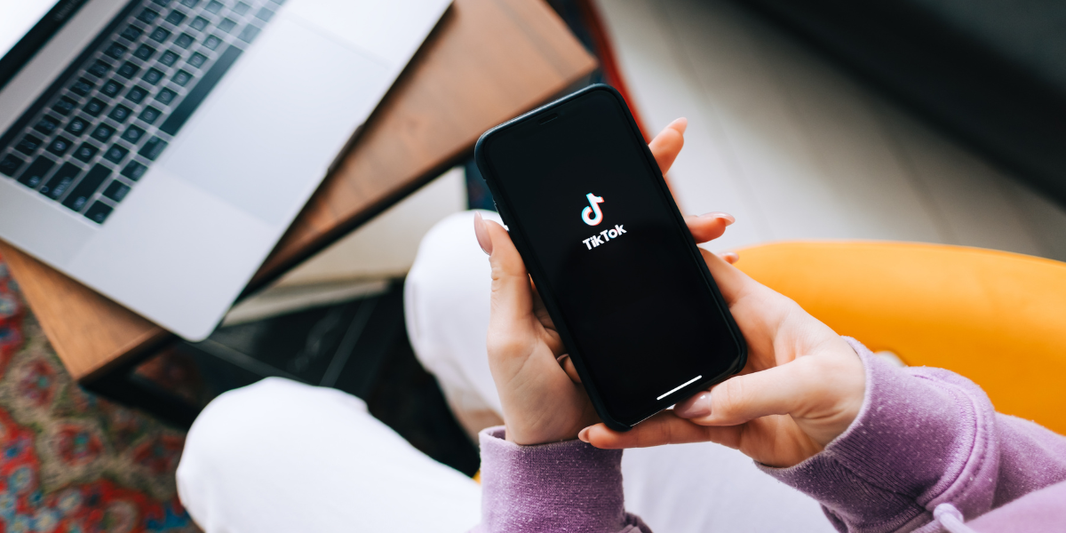 A real estate investor holds a smartphone showing the TikTok app logo, with a laptop nearby. 