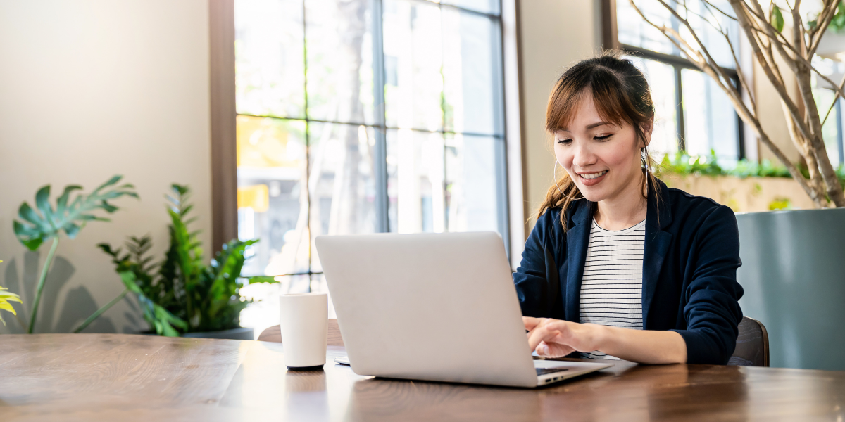 A female real estate investor working on a laptop at a wooden table in a bright café with plants.