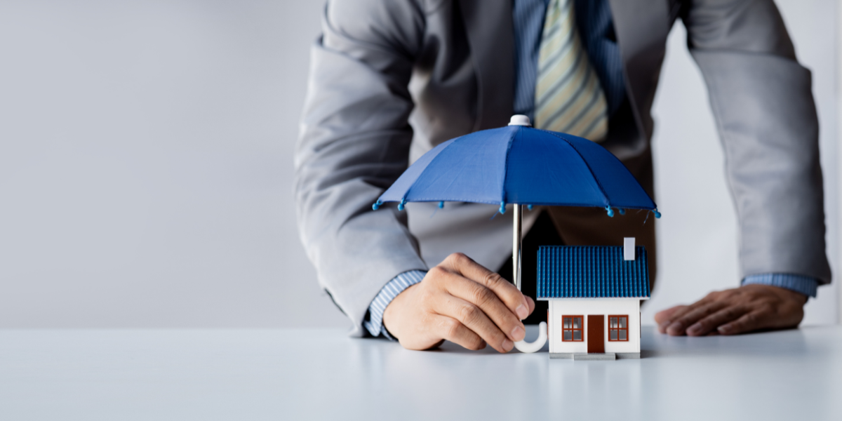 A real estate investor in a suit placing a blue umbrella over a small house model, symbolizing the protection of a rental property with rental insurance.