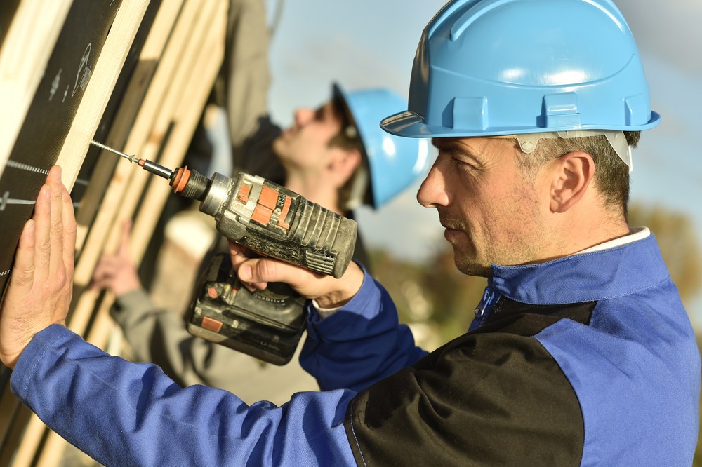 Two men in blue hardhats working with a drill on a wood wall panel for a fix and flip.
