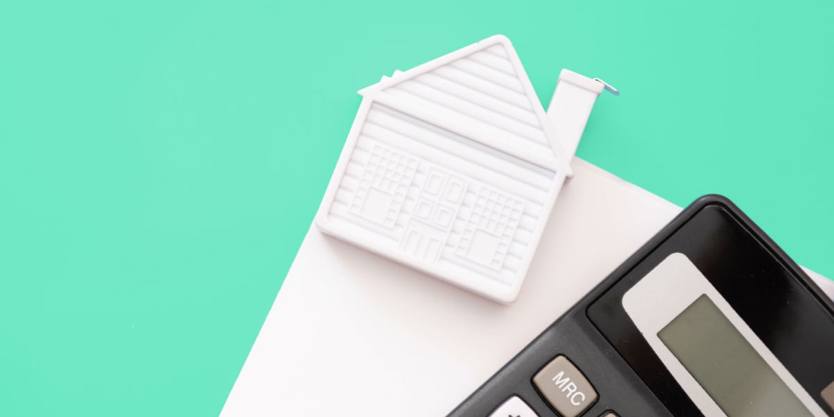 A small white house model beside a black calculator against a seafoam green and white background, symbolizing financial calculations in real estate investments 