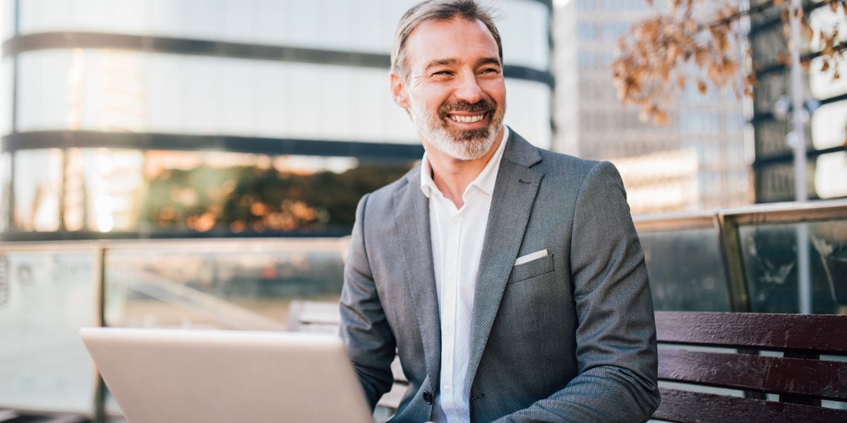 Smiling businessman using laptop outdoors with modern buildings in the background, representing successful real estate investing.