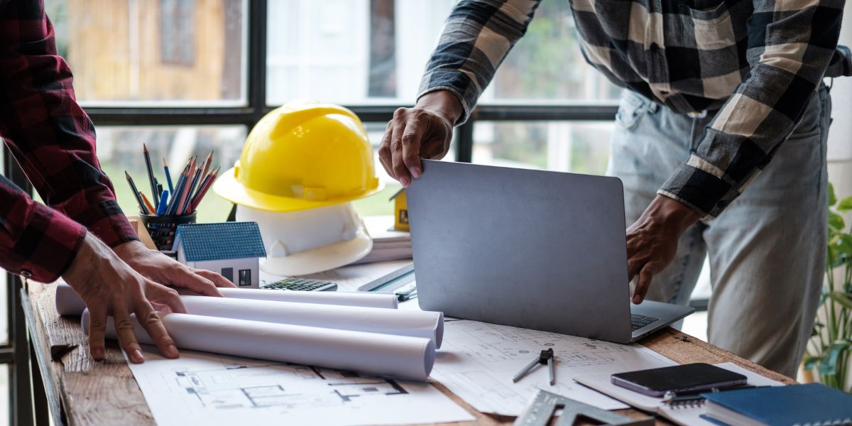 Two real estate investors discussing blueprints with a laptop and construction helmet on the table, planning an infill construction project.