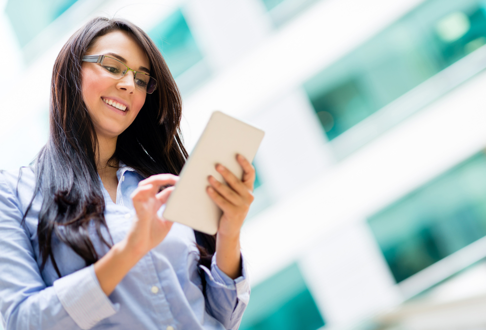 A brunette woman with glasses on a tablet smiling and looking up real estate investing apps.