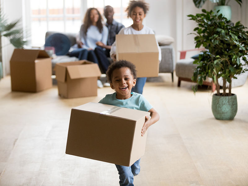 A little boy running away in a new house from a smiling family holding a moving box.