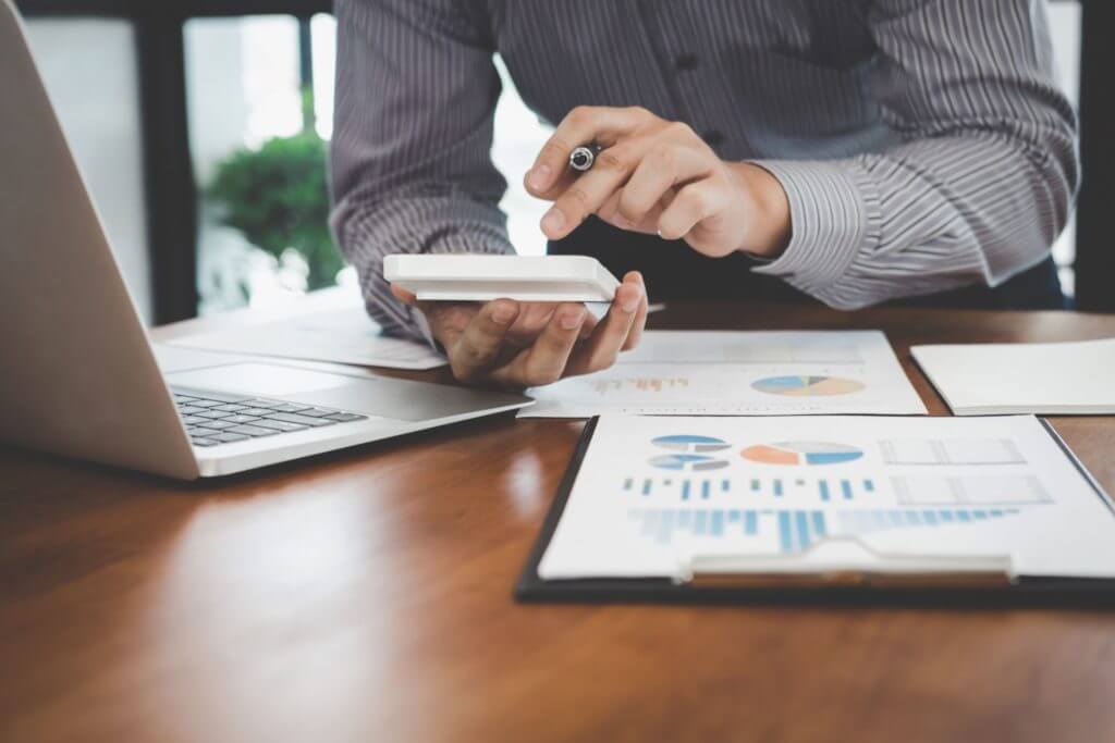 A man's hands working on a calculator with a sheet of graphs and a laptop in front of him.