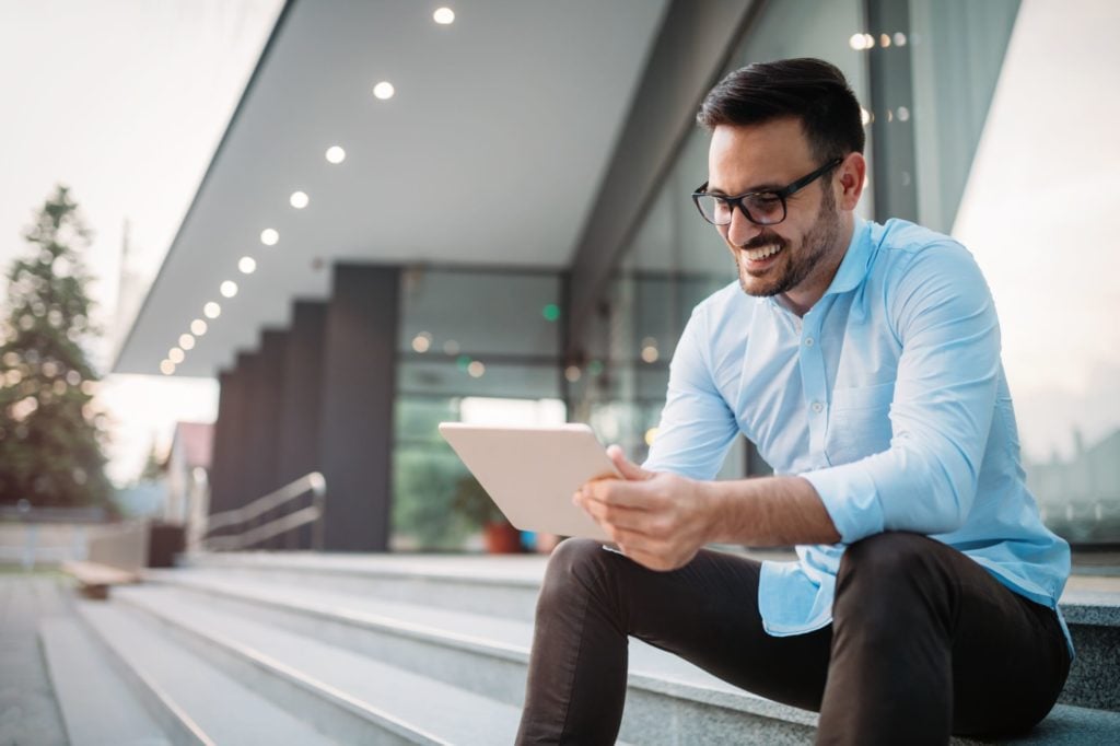 A man in glasses and a blue shirt smiling and reading a paper on the steps of an office.