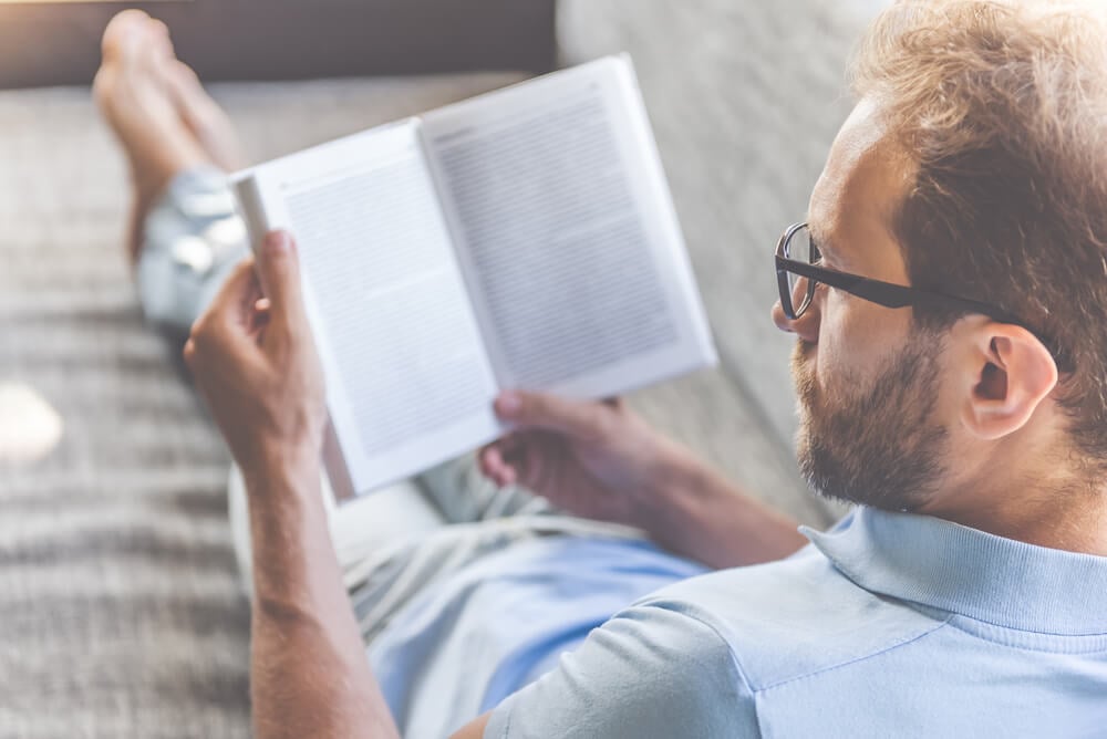 A man in a polo shirt and classes sitting on a couch reading a book.