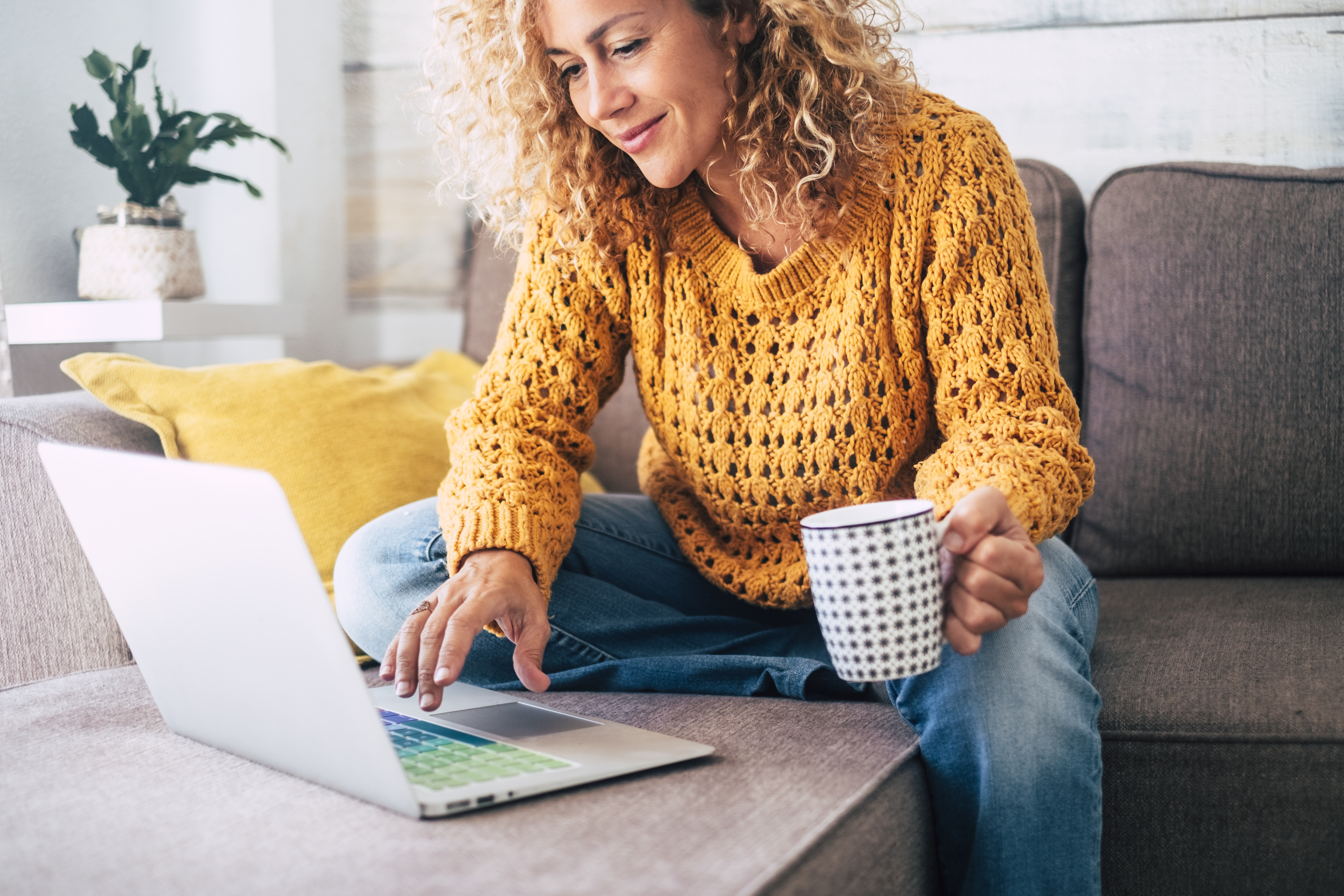 A curly-haired woman in a yellow sweater holding a coffee mug and looking at a laptop.