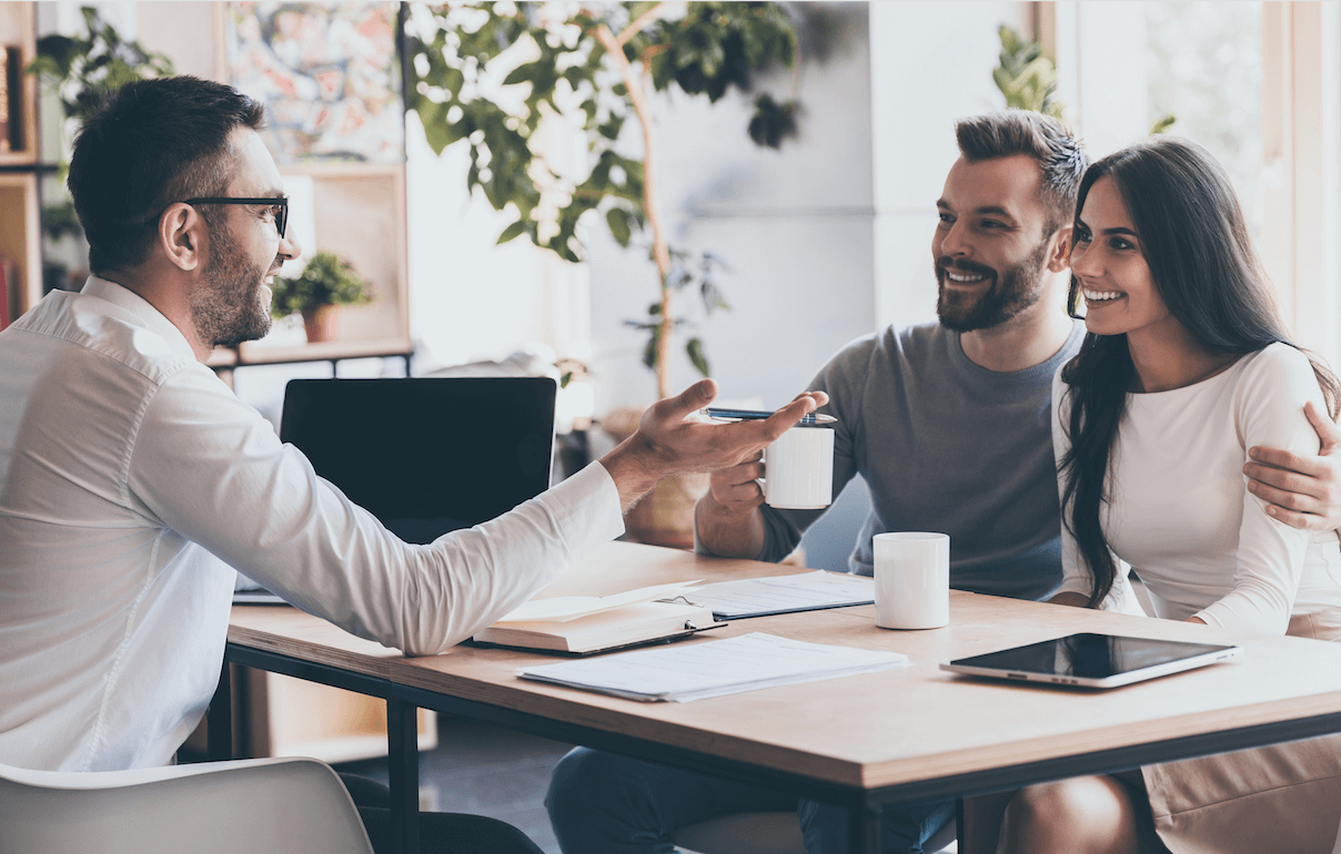 A man talking to a couple over a desk in an office with a big tree in the background.