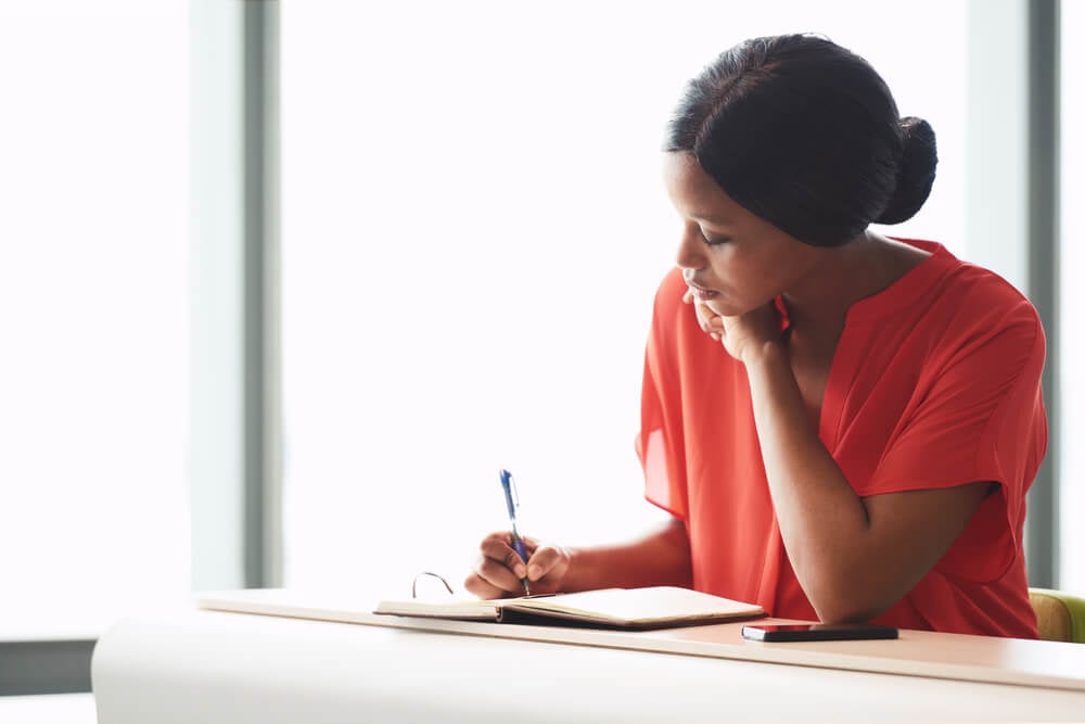 A woman in a pink shirt writing in a binder on a desk.