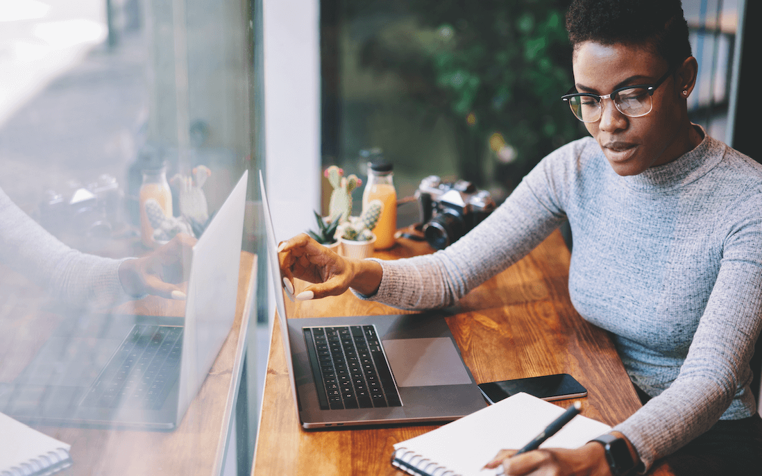 A woman in glasses on top laptop on a wooden desk next to a big window.