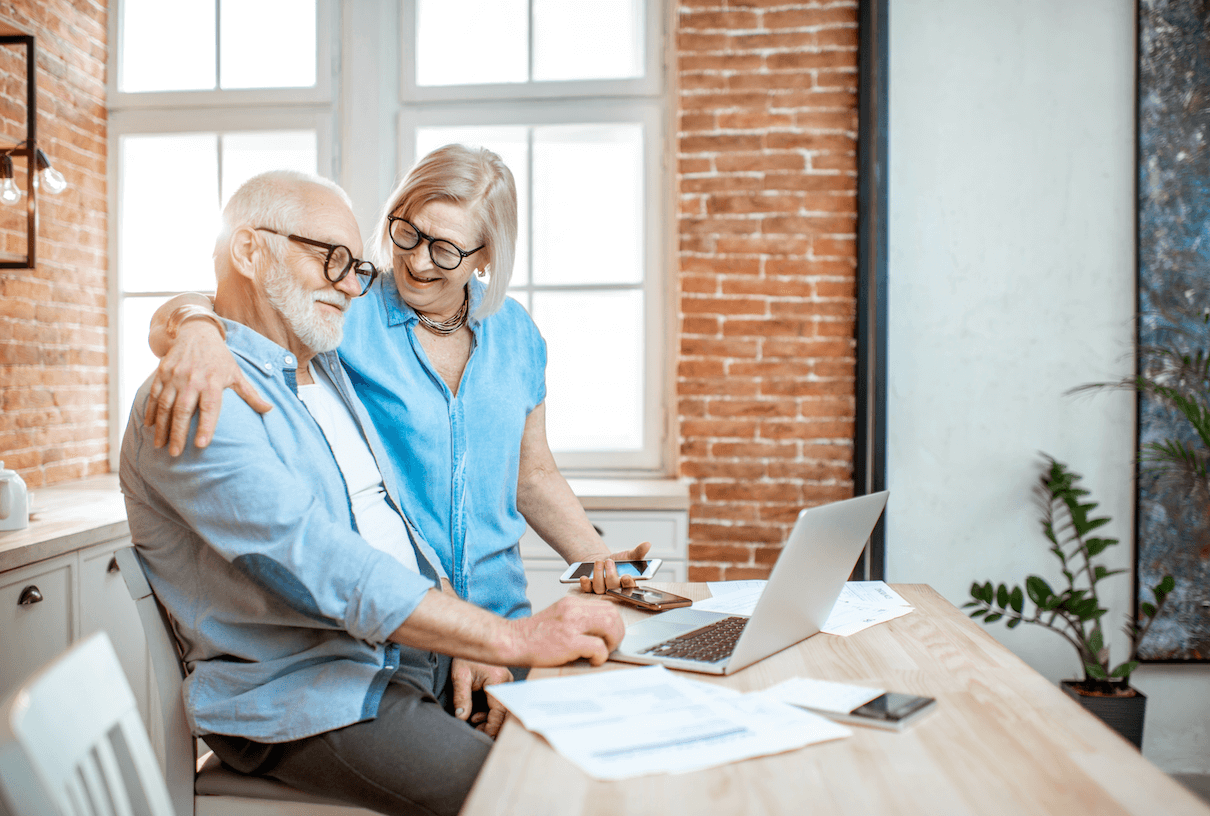 An elderly couple a desk, hugging near a laptop with a brick wall in the background.