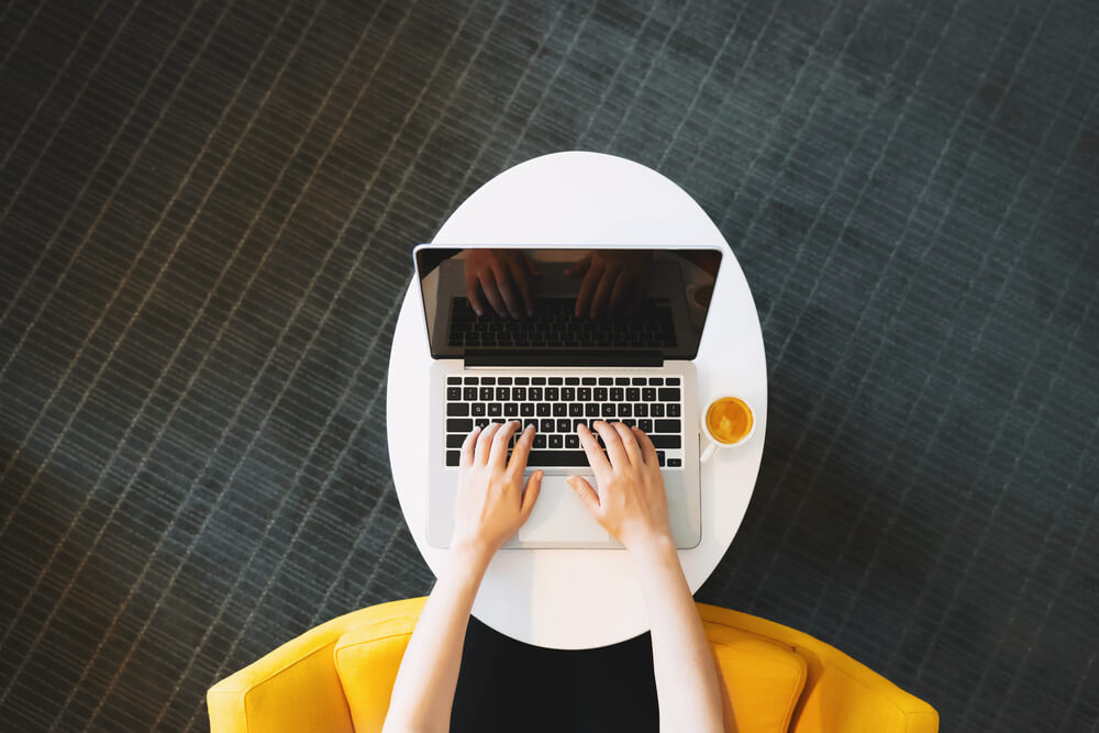 A person writing on a laptop on a white oval desk from a yellow chair.