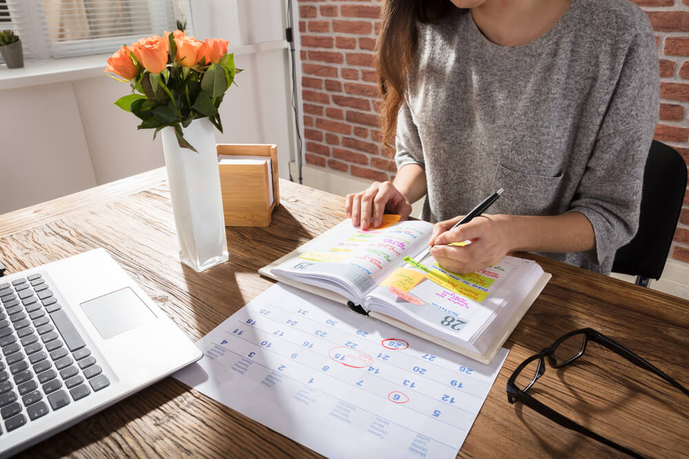 A woman working in a notebook on her wooden desk with orange flowers next to her.