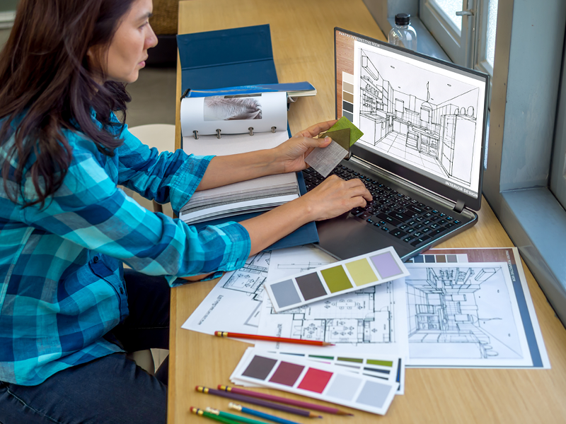 A woman in plaid looking at a house plan on a laptop computer at a desk.