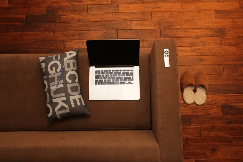 A laptop on a couch with a remote, pillow, and slippers.