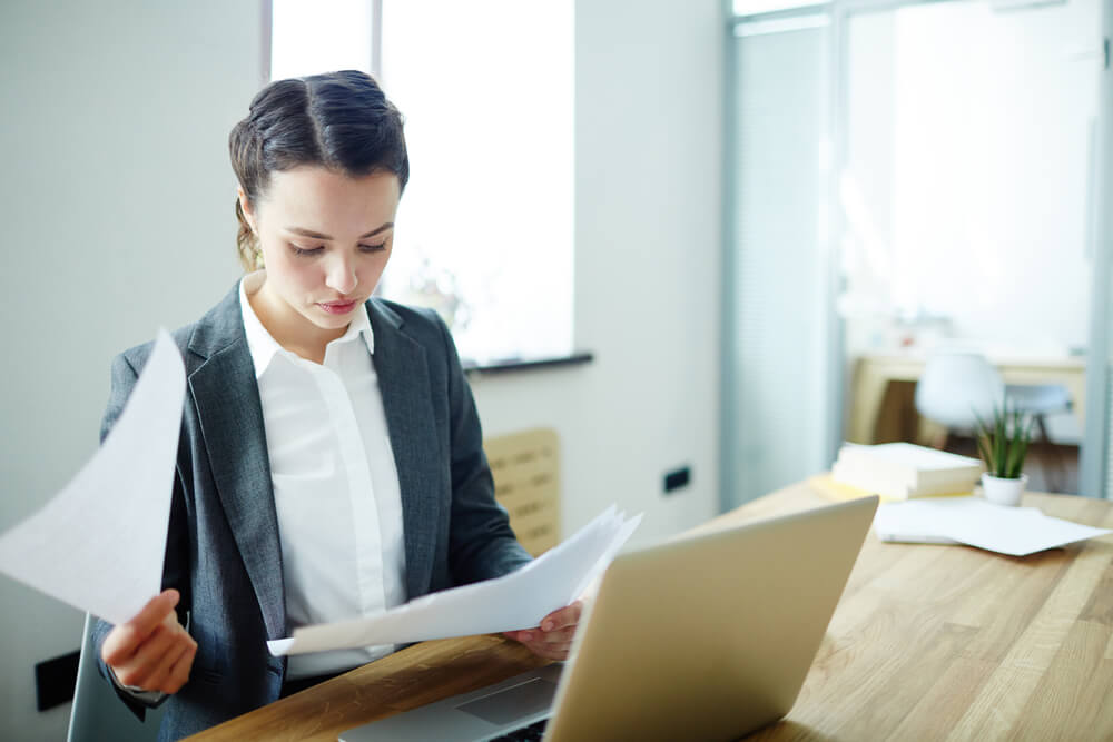 A woman at a desk reading a paper in front of a laptop.