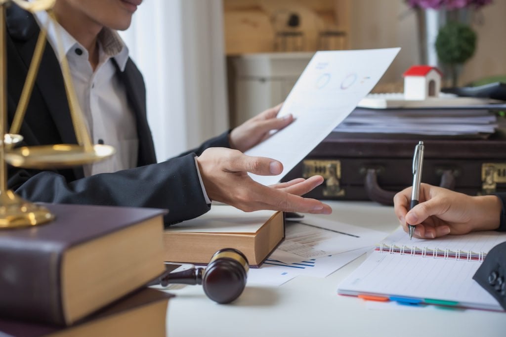 A person explaining paperwork to another person over a desk who is taking notes.