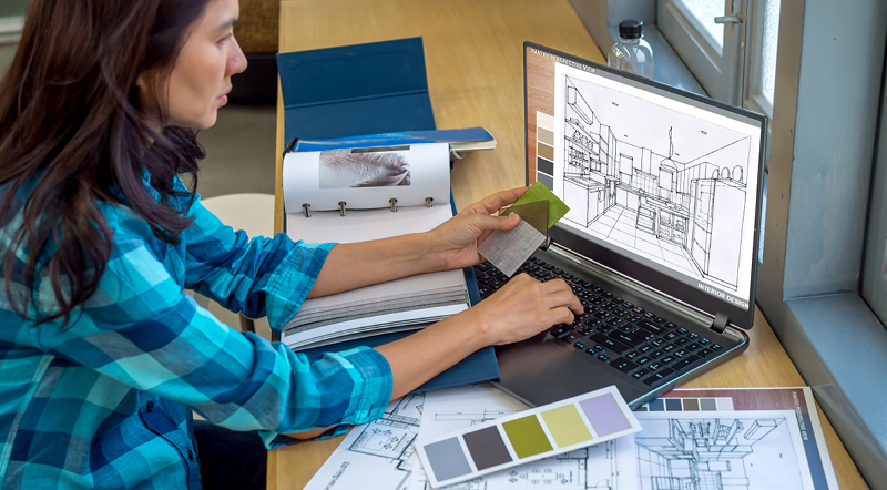A woman in a plaid shirt looking at a floor plan on a computer with paint swatches in her hand.