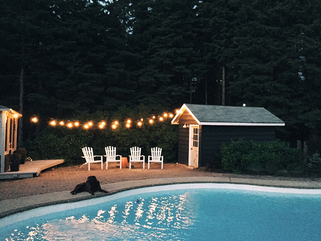 A pretty backyard with a pool, pool house, and strung lights at nighttime.