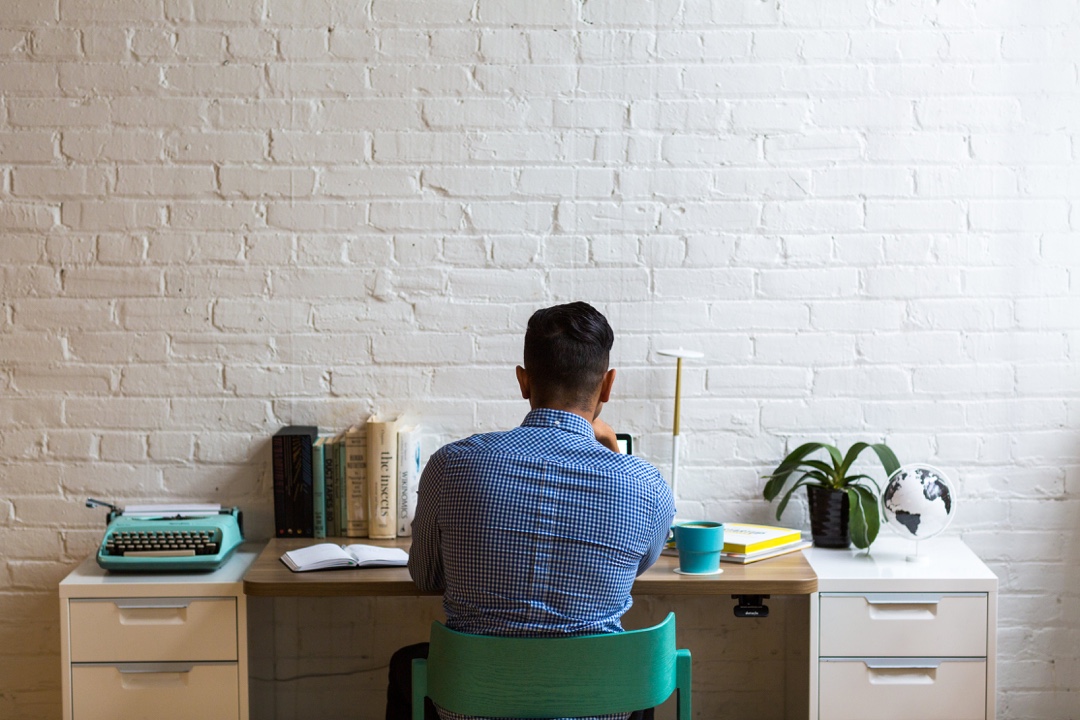 A man with his back towards the camera working at a desk in front of a white brick wall.