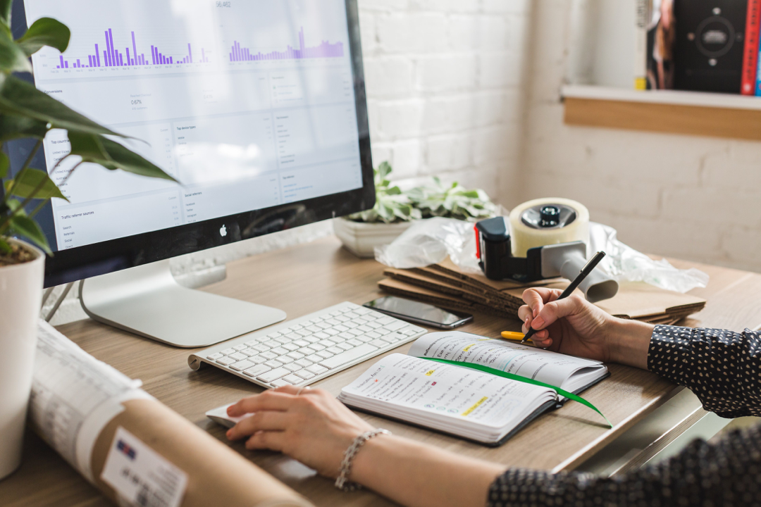 A woman's hands at a desk in front of a computer and notepad while she is finding deals.