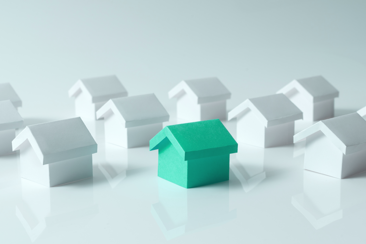 A green paper house stands out among white paper houses on a reflective surface. 