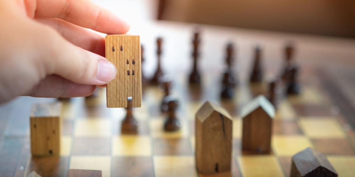 hand moving a tiny wooden house on a chess board