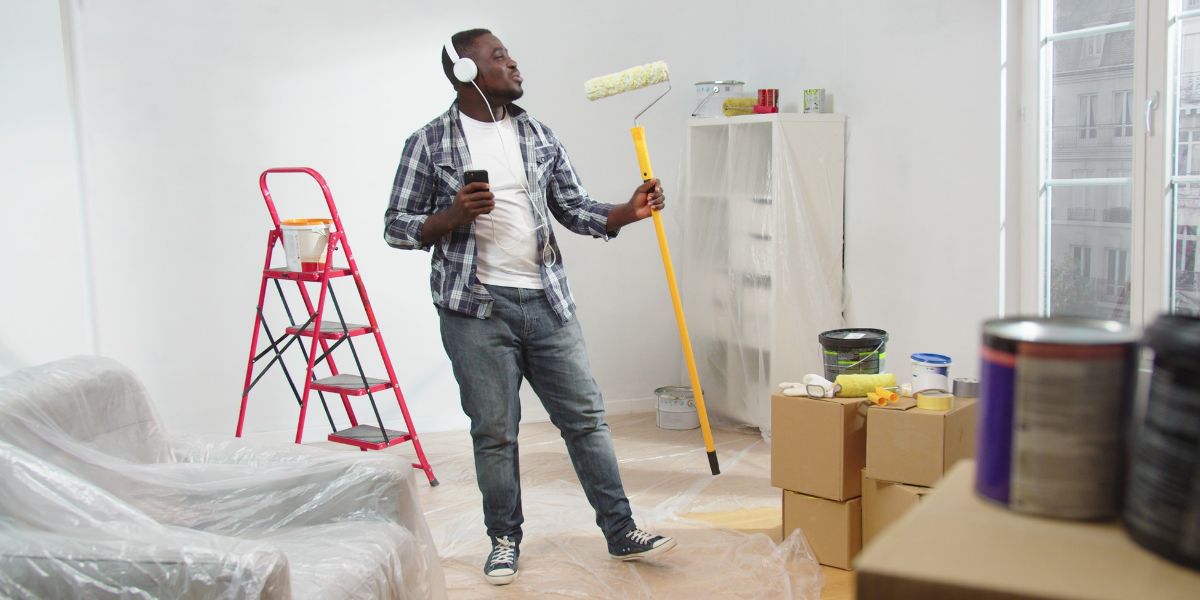 Man painting a room while enjoying music, equipped with a roller, paint cans, and a ladder, symbolizing DIY home improvement.