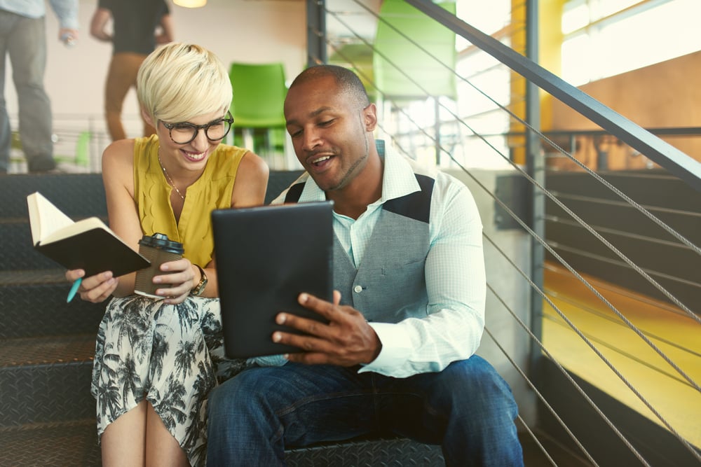 A man showing a woman something on a tablet sitting on the stairs in an office.