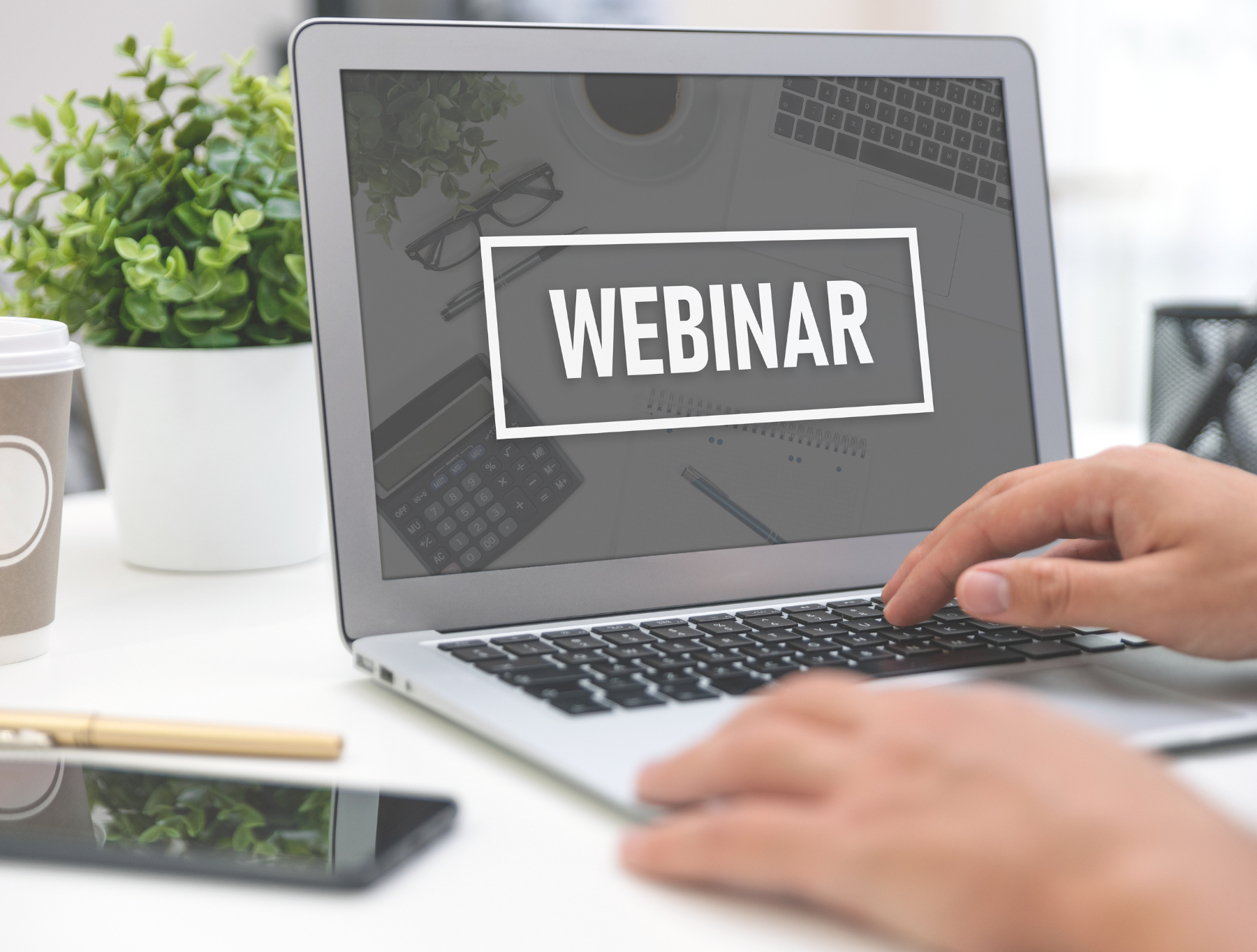 A laptop displaying the word 'WEBINAR' with a hand typing. A plant and coffee cup are in the background. 