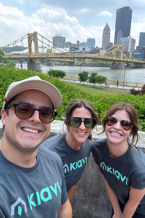 Three Kiavi employees smiling in matching Kiavi shirts, with a city skyline and bridge in the background. 