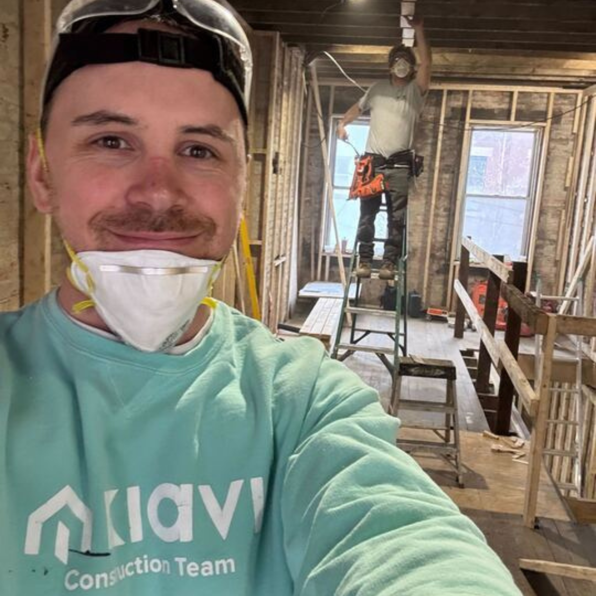 A Kiavi employee wearing a mask and helmet smiles for a selfie at a Habitat for Humanity build site, with a worker on a ladder in the background.