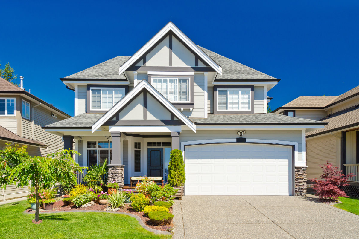 A two-story suburban home with light gray siding, a white garage door, and detailed landscaping including small trees and shrubs.