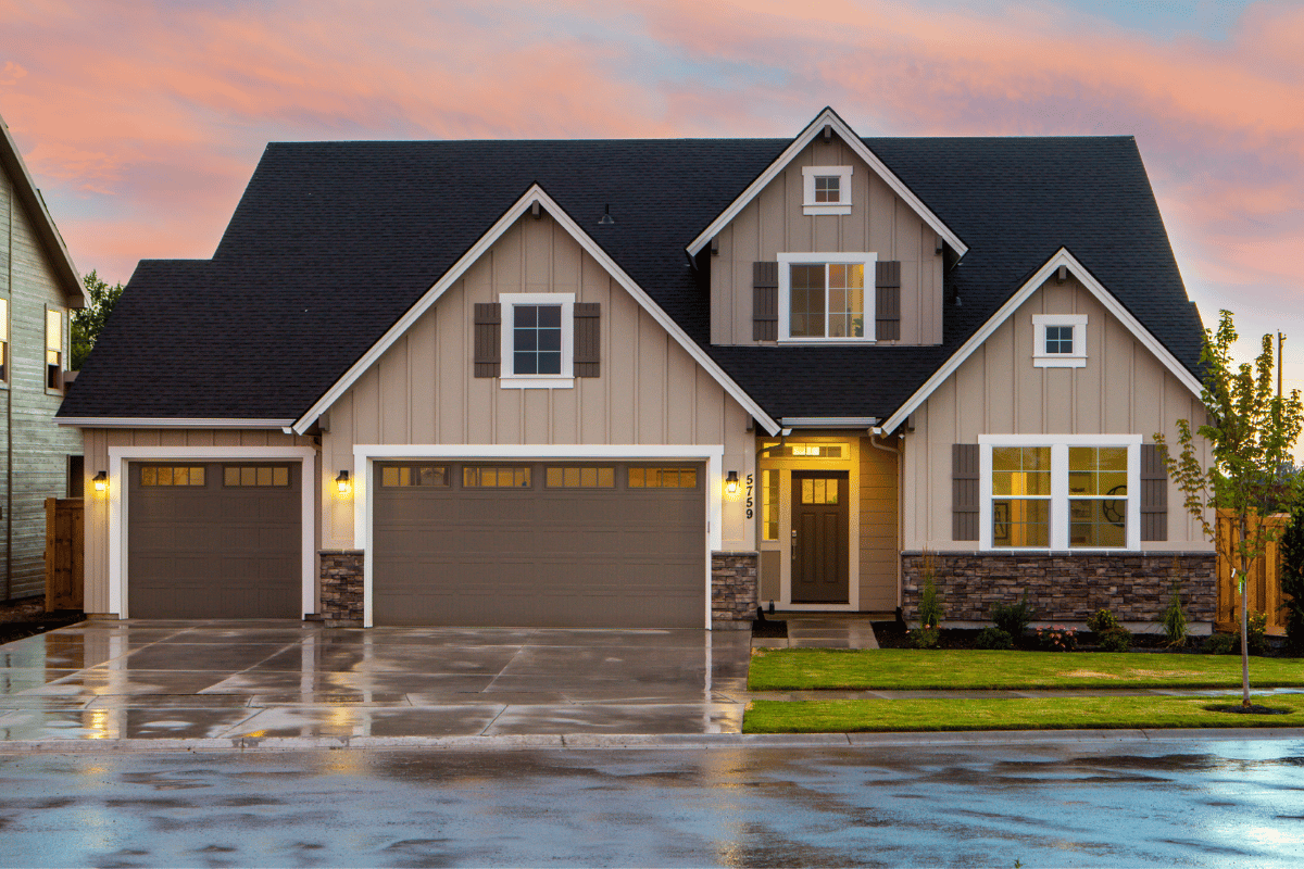 A modern two-story single-family home with beige siding, dark trim, stone accents, a three-car garage, and a wet concrete driveway beneath a sunset sky.
