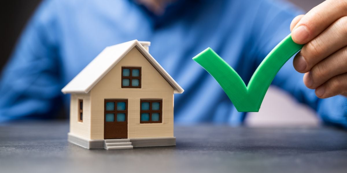 A miniature model home with a green check mark symbolizes successful approval or verification in property investment, signifying a fix-and-flip checklist.