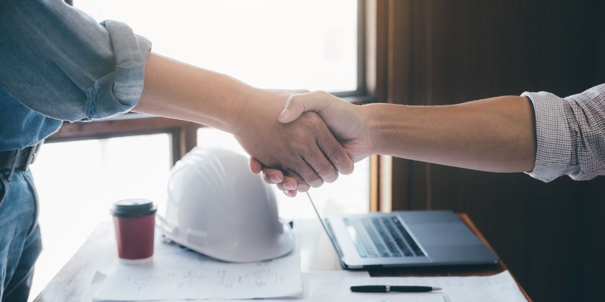 Two professionals shaking hands in agreement with construction plans and a laptop on the table, symbolizing a successful infill construction loan negotiation.