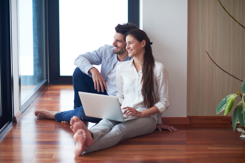 A couple sitting on the floor of a home with a laptop smiling at the distance.