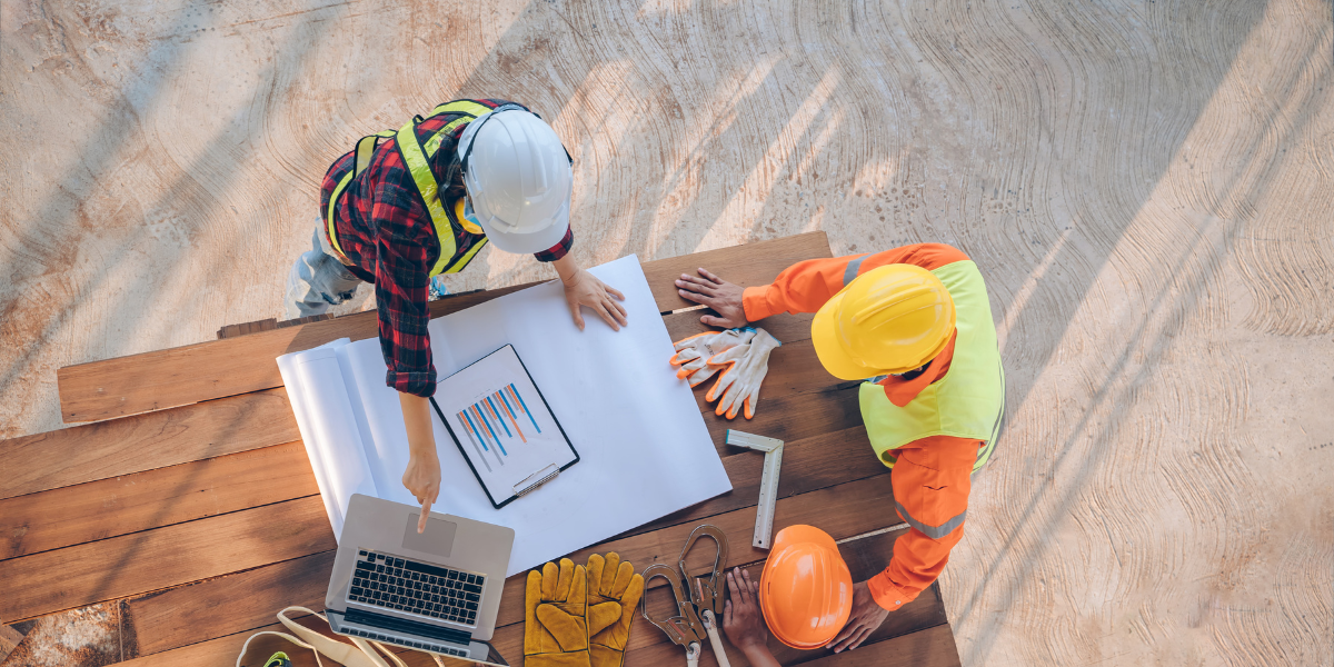 An aerial view of two ground-up construction workers in hard hats and safety vests reviewing blueprints and a laptop on a wooden table.