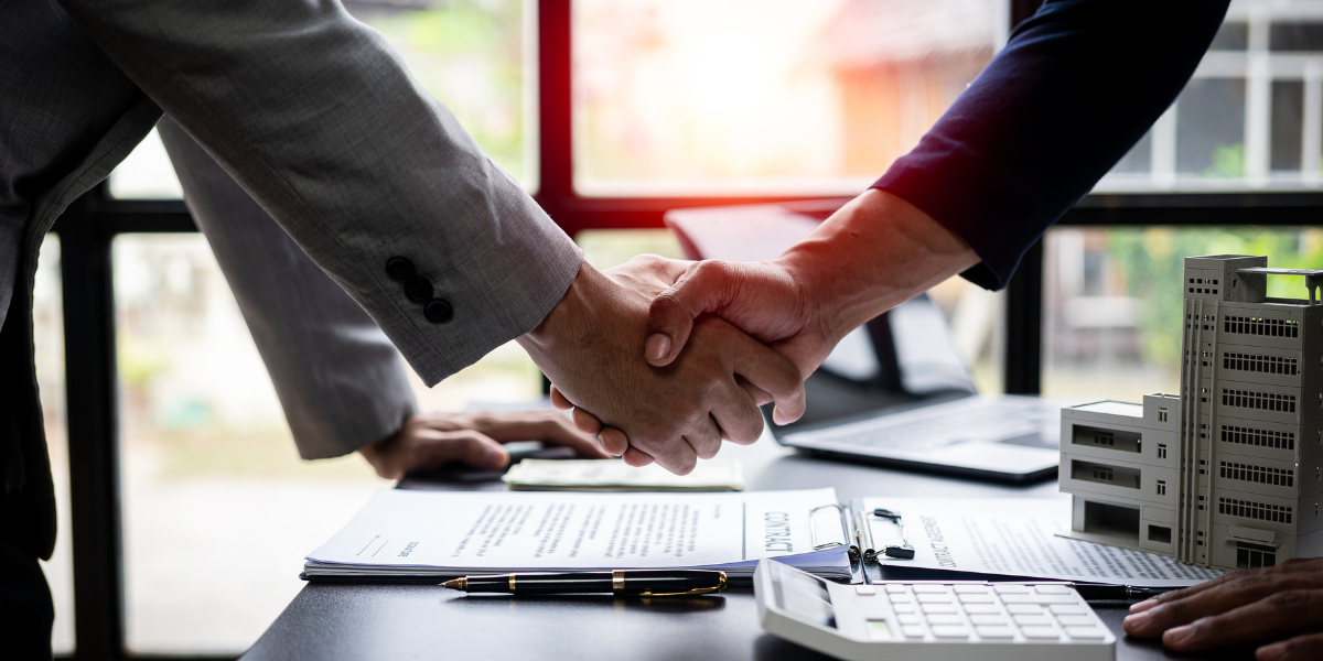 Two real estate agents shaking hands over a desk containing a signed contract, a calculator, and a 3D architectural model of a building.